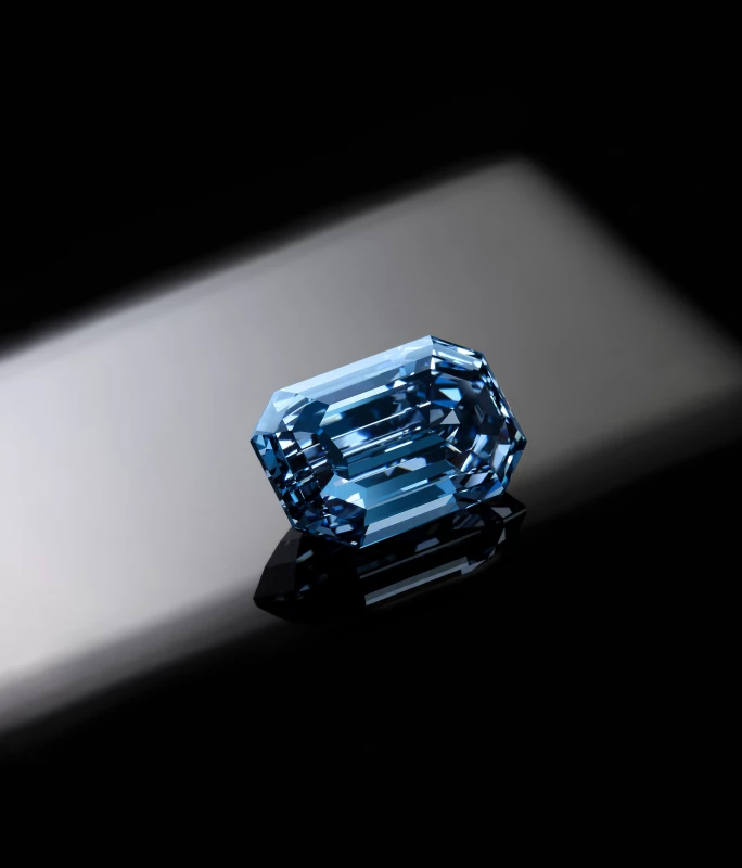 Close-up of a faceted blue gemstone with an emerald cut, resting on a reflective black surface.