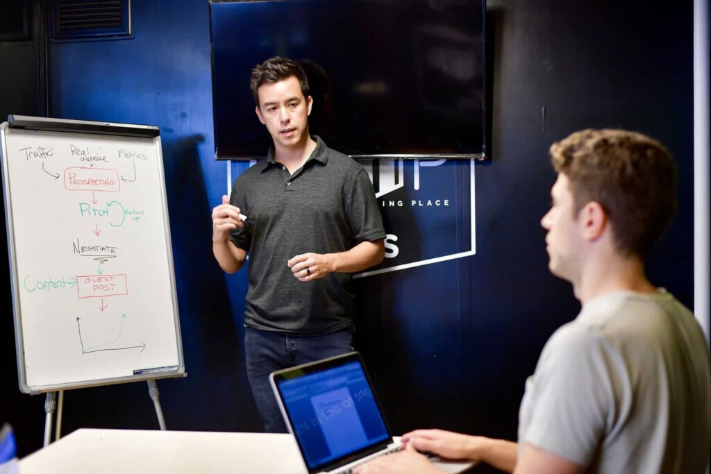 Man in a gray shirt presenting marketing strategy on a whiteboard to a colleague using a laptop in a modern office.